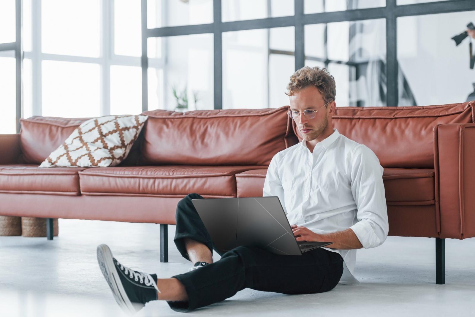 A scenario image of a man sitting on the ground with his back against a sofa, using an ASUS laptop